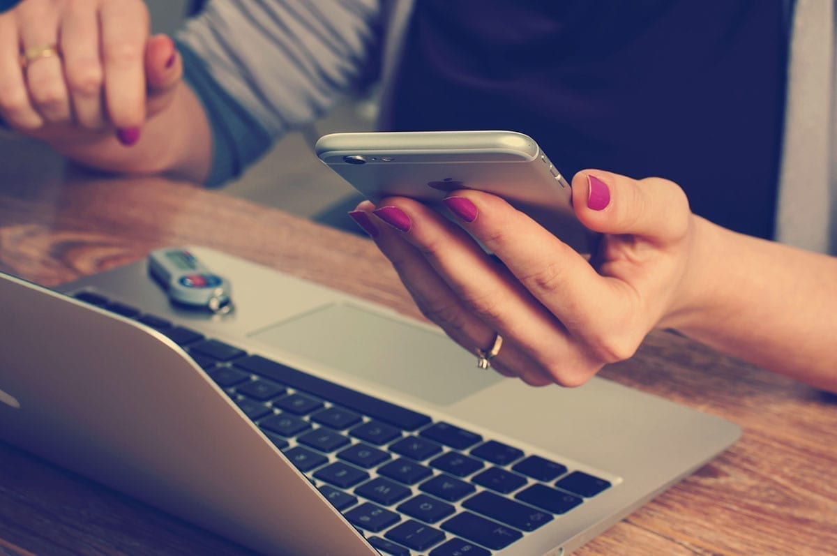 A closeup of a womans hands, one holding a smart phone and the other near a laptop computer; highlighting assistance programs available for missed mortgage payments and the need to reach out early