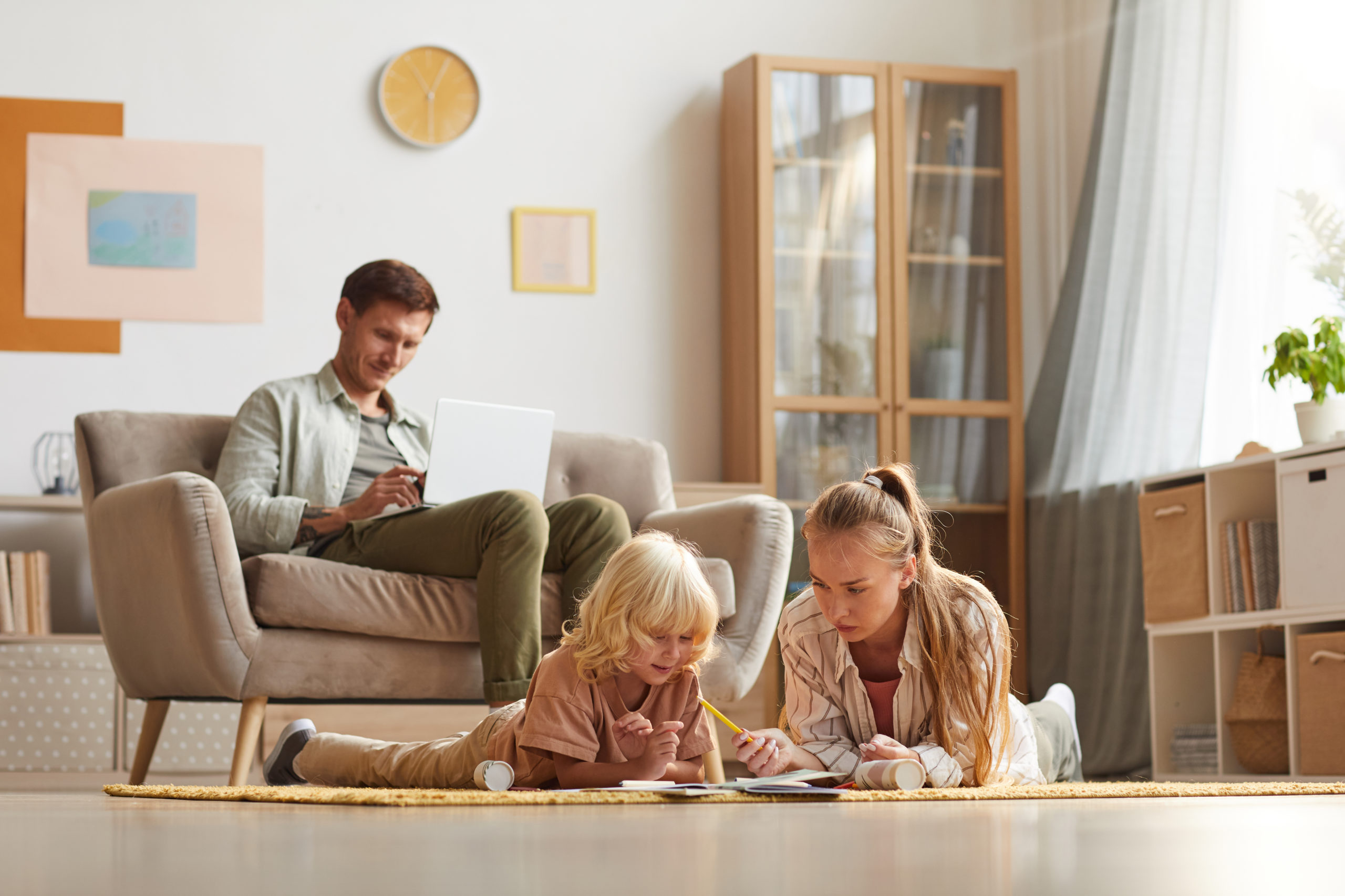 Mother with her son lying on the floor and drawing while their father working on laptop sitting on sofa they are in the living room