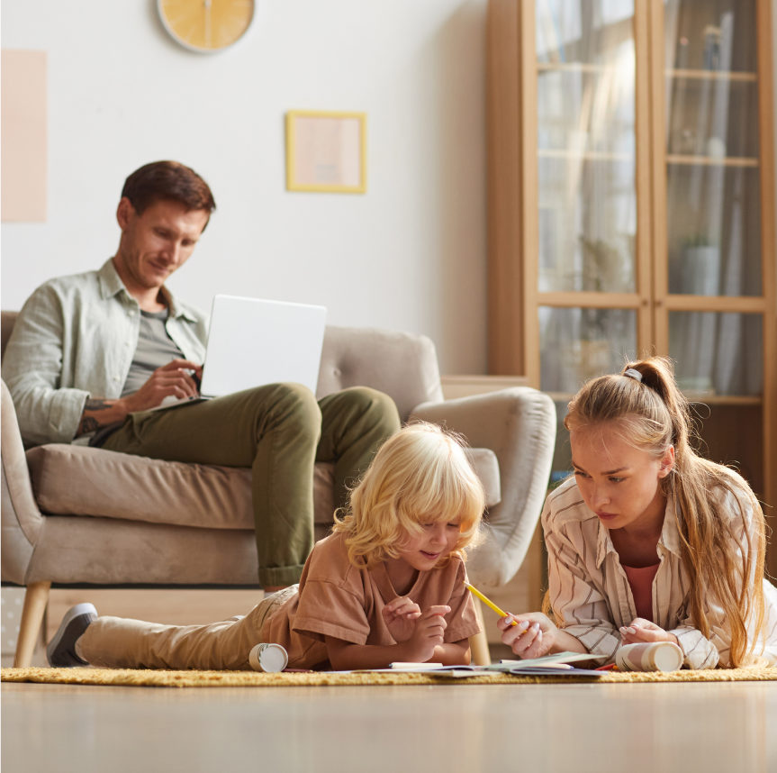 A young family in their living room, with the father on the couch looking at his computer, and the mother and child on the floor playing with papers and a pencil