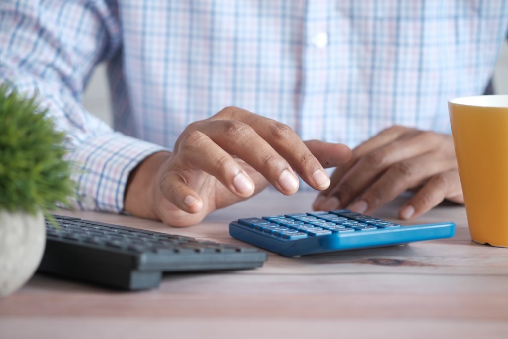 closeup of a man's hands using a calculator to determine whether he can afford a mortgage in BC, with a desk plant and coffee cup in the foreground